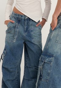 Bredbyxiga cargobyxor i urtvättad blå denim med flera fickor, distinkt sömnad och texturerad tyg. Har en lös passform och användardesign.