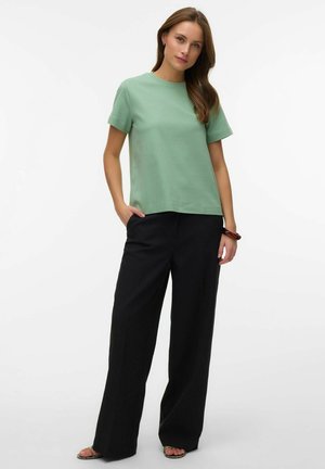 Kvinde stående med hænderne i lommerne på sorte bukser med vide ben, iført en lysegrøn kortærmet top og sandaler, mod en hvid baggrund.