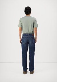 Green crew-neck t-shirt and dark blue straight-cut jeans with visible stitching and a brown leather back patch, worn with light brown shoes.
