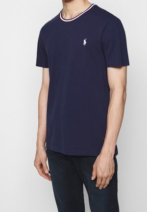 Uomo che indossa una maglietta a maniche corte blu navy con rifiniture bianche e rosse sul colletto e un piccolo logo bianco sul petto a sinistra.