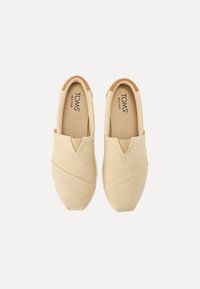 Des chaussures slip-on beiges avec une texture en chevron et des accents marron. Présentent un design en V élastiqué et une semelle en caoutchouc blanche.