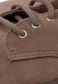 Brown suede shoe close-up with braided laces, metal eyelets, stitching detail, and a smooth texture.