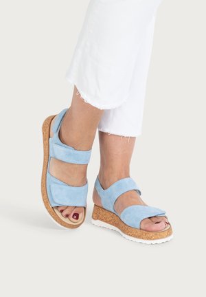 Füße, die hellblaue Wildleder-Plattform-Sandalen mit Korksohlen und weißen Unterseiten tragen, kombiniert mit weißen Hosen mit ausgefranstem Saum.
