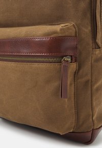 Hnědá plátěná taška s koženou kapsou na zip a zdobením z hnědé kůže na spodní části. Texturovaná látka s pevnou konstrukcí.