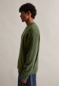 Pulover verde tricotat, cu guler rotund și mâneci lungi, realizat dintr-un material texturat moale. Purtați cu blugi albaștri, prezentat din lateral.