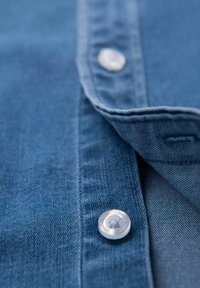 Denimhemd mit blauer Färbung, ausgestattet mit einem Knopfverschluss aus klarem Kunststoff und einem genähten Kragen mit sichtbarer Textur und Nähten.