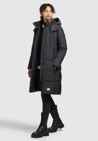 Black padded long coat with a hood, featuring zip and button closure, vertical seams, and a logo patch on the lower front. Worn over black clothing and boots.
