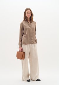 Blusa beige in satin con bottoni frontali, abbinata a pantaloni larghi in lino beige chiaro e una borsa strutturata in pelle marrone. Scarpe nere a punta.