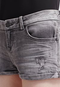 Shorts de mezclilla grises con un parche desgastado en el frente, que presentan un diseño clásico de cinco bolsillos y un dobladillo enrollado en la parte inferior.