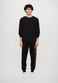 Felpa nera con tasca frontale, abbinata a pantaloni della tuta neri con un logo dorato verticale. Indossata con sneakers bianche.