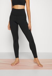 Korkeavyötäröiset mustat leggingsit, joissa on sileä pinta, sivusaumat ja tiukka muotoilu, esitelty vaaleanpunaisella taustalla.