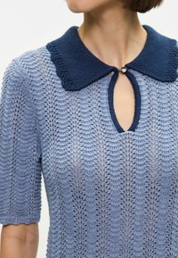 Ανοιχτό μπλε πλεκτό τοπ με υφή σε σχέδιο φύλλου, που διαθέτει σκούρο μπλε γιακά, άνοιγμα σε σχήμα κλειδώματος και κοντά μανίκια με οδοντωτές άκρες.