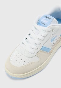 Biele kožené tenisky s svetlo modrými akcentmi, zamšovým prekrytím, perforovanou špičkou a bielymi šnúrkami. Logo Lacoste na boku a jazyku.