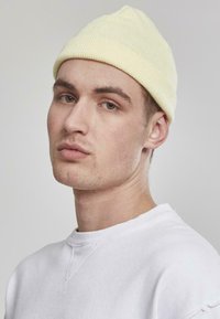 Beanie tricotat galben cu o potrivire strânsă; purtat cu un hanorac alb simplu. Fundalul este neutru și neintruziv.