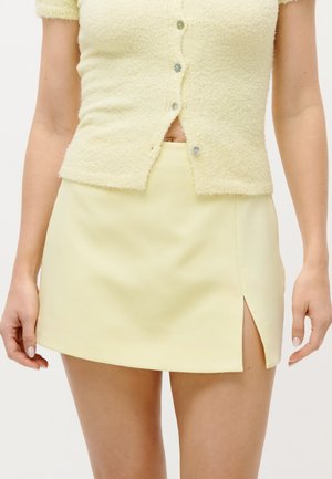 Sieviete, kas valkā gaiši dzeltenu kardiganu ar pogām un atbilstošu mini svārku ar atvērumu priekšpusē, stāv uz vienkārša fona.