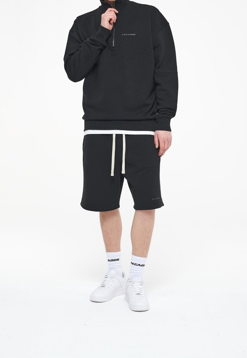 Homme portant un sweat-shirt noir à fermeture éclair quart de zip, un short noir avec cordon de serrage, des chaussettes blanches avec logo, et des baskets blanches, debout devant un fond blanc.