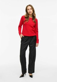 Un pull rouge texturé à manches longues, associé à un pantalon large noir taille haute et des escarpins noirs à bout pointu.