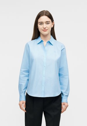 Mladá žena s rovnými hnedými vlasmi, oblečená v svetlomodrej košeli na gombíky a čiernych nohaviciach, stojaca pred jednofarebným bielym pozadím.