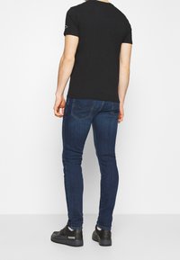 Schwarzes T-Shirt mit kurzen Ärmeln kombiniert mit dunkelblauen Slim-Fit-Jeans und schwarzen Sneakers. Sichtbares Logo auf dem T-Shirt-Ärmel, dezente Nahtdetails an der Jeans.