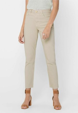 Vrouw draagt lichtbeige slim-fit broek met rafelige zomen en bruine enkelriem blokhak sandalen, staand tegen een effen achtergrond.