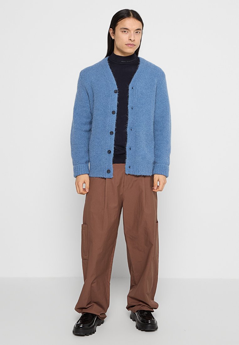 Blau gestrickte Strickjacke mit V-Ausschnitt, Knopfschluss und gerippten Bündchen. Getragen über einem schwarzen Rollkragenpullover und kombiniert mit braunen Cargo-Hosen.