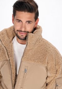 Beige zip-up jakke med en luksuriøs, tekstureret fleece-overdel og en glat underdel. Har sølvfarvet lynlås og sidelommer.