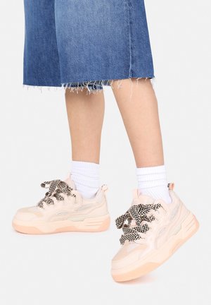 Chunky sneakers in zacht roze en crème, met een gestructureerd mesh en een zigzag patroon in de veters. Vergezeld door witte geribde sokken en denim.