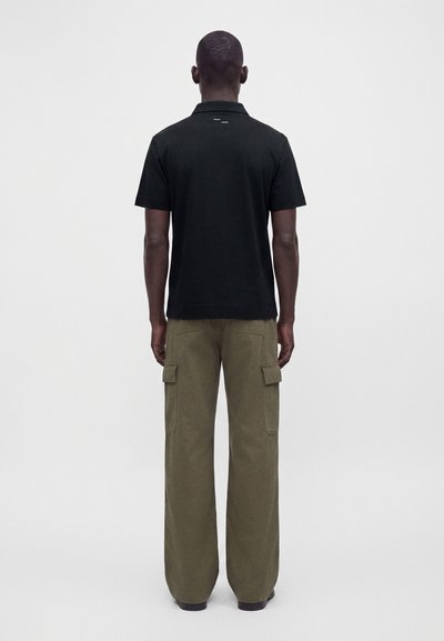 Homme debout de dos, portant une chemise noire à manches courtes et un pantalon cargo ample vert olive avec des poches avant.