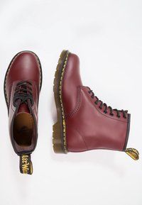 Paire de bottines en cuir bordeaux à lacets avec surpiqûres jaunes, semelles épaisses translucides, et boucles de talon noires et jaunes avec logo, présentées sur fond blanc.