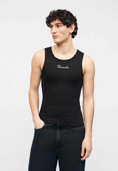 Jeune homme aux cheveux bouclés portant un débardeur noir avec le texte "Endorphin", jean foncé, regardant sur le côté avec une main dans la poche.