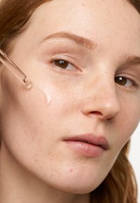 Sérum clair appliqué sur la peau à l'aide d'un compte-gouttes. La peau est claire et lisse avec de petites taches de rousseur, et le sérum crée un léger éclat.