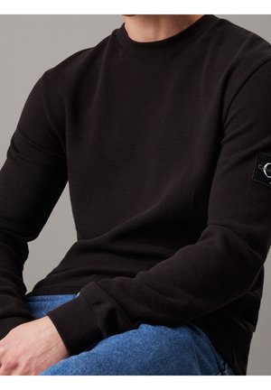 Mężczyzna ubrany w czarny, teksturowany sweter z długim rękawem z naszywką z logo Calvin Klein na rękawie, w połączeniu z niebieskimi dżinsami, siedzący z rękami spoczywającymi.