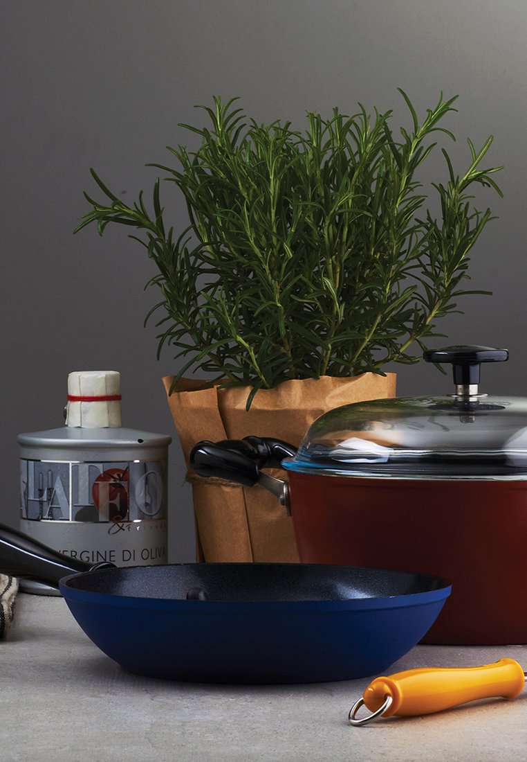 Padella blu, pentola rossa con coperchio di vetro, lattina d'olio d'oliva, sacchetto di carta marrone con erbe, e uno strumento con manico arancione su una superficie grigia.