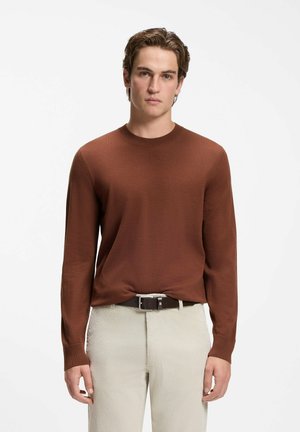 Giovane uomo con maglione marrone a maniche lunghe e pantaloni beige, in piedi di fronte a uno sfondo chiaro e uniforme.