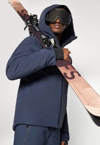 Casaco de esqui azul-marinho com capuz, usado sobre uma camada térmica preta. Um esqui com um design rosa e castanho está pendurado sobre o ombro.