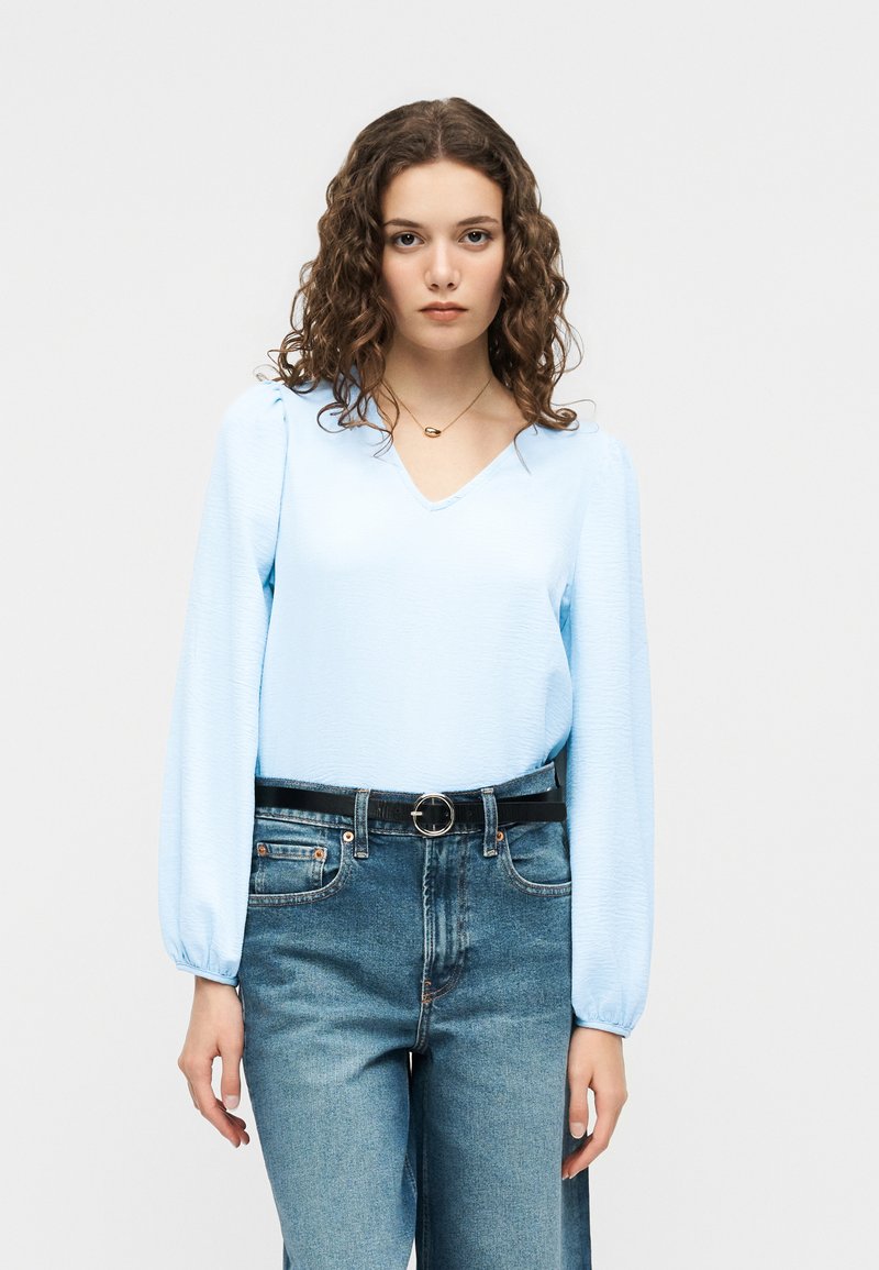 Γυναίκα με σγουρά μαλλιά που φοράει ανοιχτό μπλε μπλούζα με μακριά μανίκια, ψηλόμεσα μπλε τζιν και μαύρη ζώνη με στρογγυλή αγκράφα.