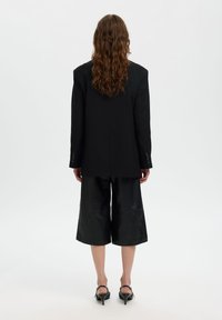 Άτομο με μακριά κυματιστά μαλλιά φοράει μαύρο υπερμεγέθες σακάκι, μαύρο φαρδύ παντελόνι cropped και μαύρα παπούτσια με χαμηλό τακούνι, στέκεται με την πλάτη προς τα εμάς.