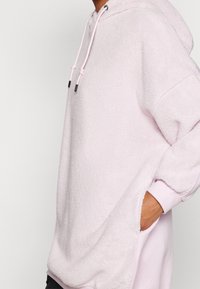 Felpa cappuccio rosa pelosa con tasca a marsupio, collo con cordino e vestibilità comoda. Presenta polsini a coste e una texture morbida.