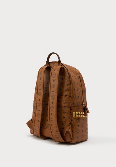 Brown leather backpack with black MCM logos, two padded shoulder straps, gold zipper, and gold stud detail on side pocket, angled view.
