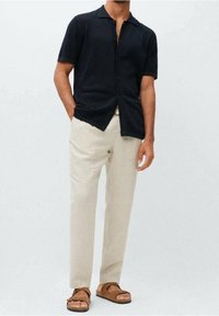 Zwarte korte mouwen polo, lichte stof, knoopsluiting aan de voorkant, gecombineerd met beige linnen broeken en bruine sandalen. Neutrale kleuren.