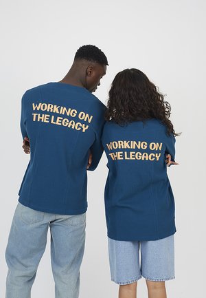 Dwie osoby ubrane w niebieskie koszulki z żółtym napisem „WORKING ON THE LEGACY” na plecach, stojące obok siebie tyłem.