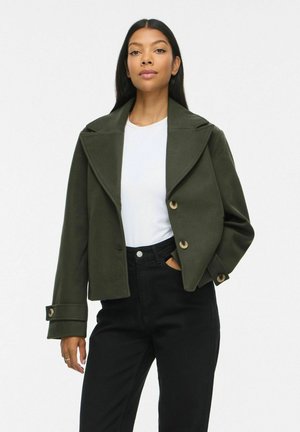 Donna che indossa una giacca verde scuro con bottoni sopra una camicia bianca e pantaloni neri, in piedi con una mano in tasca su uno sfondo semplice.
