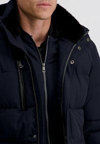 Man in een donkerblauwe winterjas met zichtbare metalen ritsen en drukknoopjes, voorzien van een zwarte gevoerde kraag van bont.