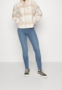 Sudadera de forro polar a cuadros en beige y blanco, vaqueros ajustados azules y zapatillas negras y blancas con un patrón texturizado.
