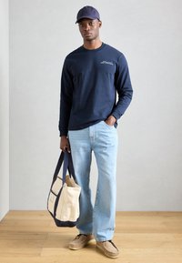 Tumesinine pika varrukaga särk, helesinised teksad, beež kott tumedate aktsentidega ja pruunid libisemisjalanõud. Neutraalne taust, puidust põrand.