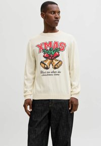 Maglione a maglia crema con testo rosso "XMAS", design con agrifoglio e grafica delle campane. Maniche e orlo a coste con vestibilità rilassata. Abbinato a jeans scuri.