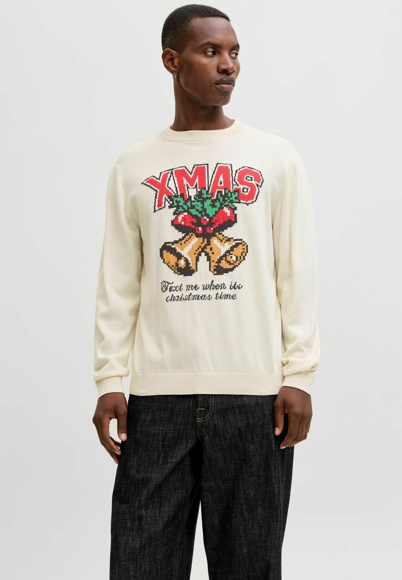 Maglione a maglia crema con testo rosso "XMAS", design con agrifoglio e grafica delle campane. Maniche e orlo a coste con vestibilità rilassata. Abbinato a jeans scuri.