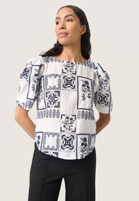Vit blus med marinblå blommönster, kort puffärmar och rund halsringning, tillsammans med svarta byxor. Mjuk, strukturerad tyg.