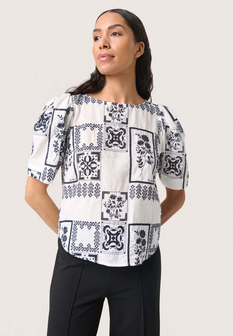 Blusa bianca con motivi floreali blu navy, maniche corte a sbuffo e scollatura rotonda, abbinata a pantaloni neri. Tessuto morbido e strutturato.