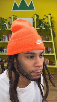 Căciulă portocalie tricotată cu logo alb Nike, purtată de o persoană cu păr lung, negru, în dreadlocks. Fundalul prezintă un perete galben strălucitor și rafturi verzi.